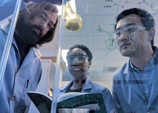 Three chemists standing in front of a fume hood, all dressed in blue lab coats and wearing safety glasses. One is holding a round-bottom flask of a yellow compound while the other two look on